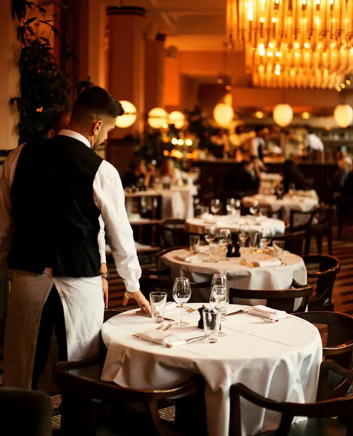 Cecconi’s Los Angeles: Waiter setting tables in a warmly lit restaurant interior.